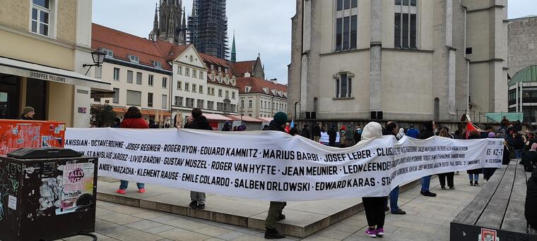 Eine Kundgebung auf dem Regensburger Neupfarrplatz mit einem langen, weißen Banner, auf dem die Namen von Opfern des NS-Regimes geschrieben sind.