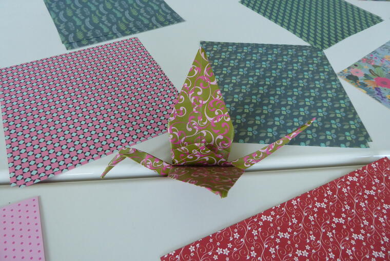 Ein Papierkranich mit buntem Muster auf einem Tisch mit verschieden farbigen Origamipapieren