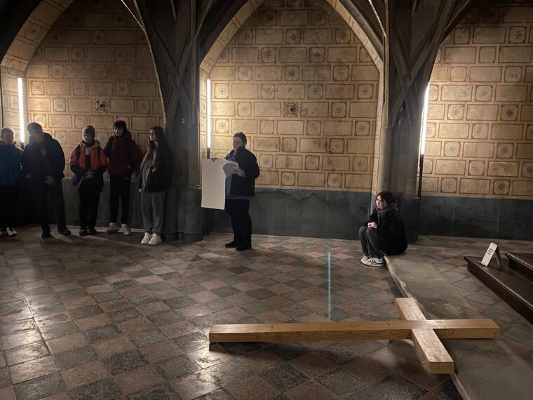 Menschen sitzen und stehen in einer Kapelle. Vor dem Altar liegt ein großes Holzkreuz am Boden.