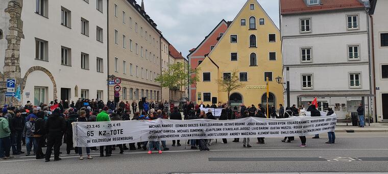 Viele Menschen bei einer Gedenkveranstaltung, ein weißes Banner mit den Namen von NS-Opfern wird gezeigt.